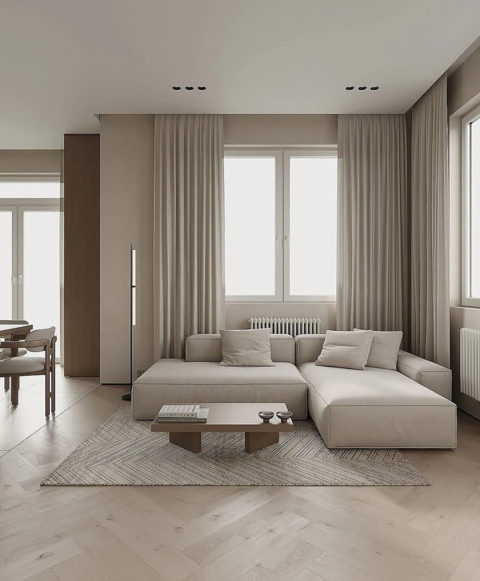 A pale sectional sofa, compact table, textured rug and layered curtains create a quiet lounge composition.